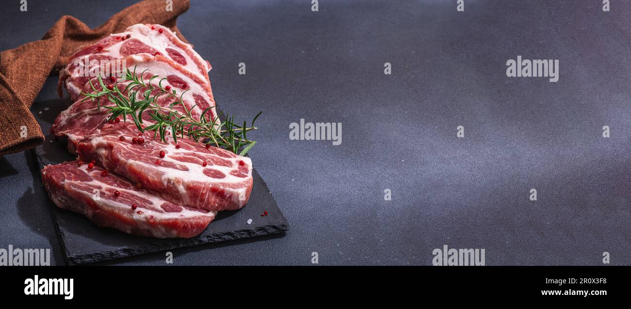 Rohes Schweinenackfleisch. Hacksteak, roter Pfefferkörner, Knoblauchzehen, Meersalz und frischer Rosmarin. Trendiger Standfuß, schwarzer Hintergrund, flach liegend, Bannerformat Stockfoto