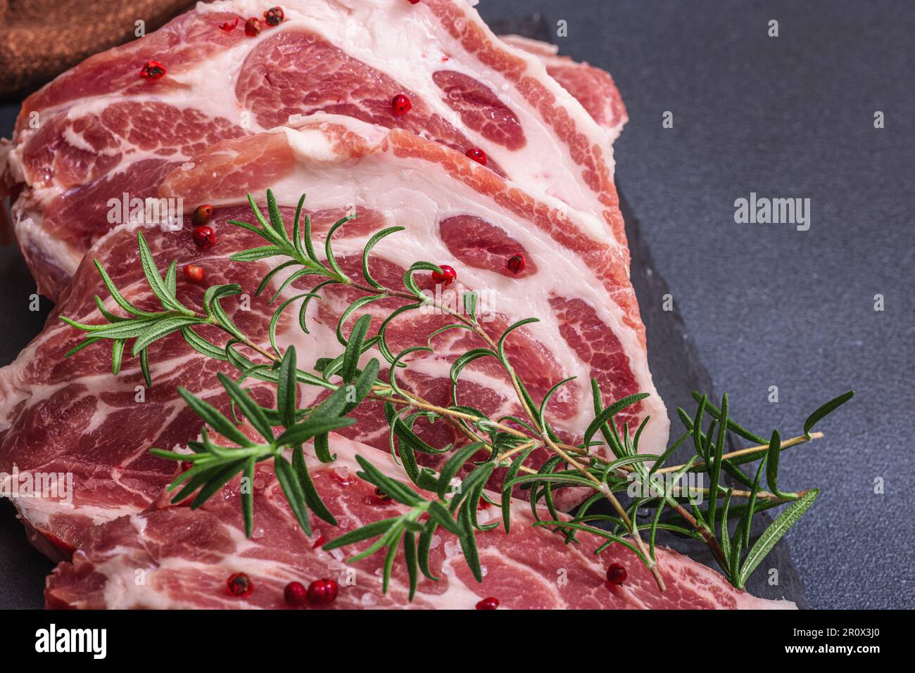 Rohes Schweinenackfleisch. Hacksteak, roter Pfefferkörner, Knoblauchzehen, Meersalz und frischer Rosmarin. Trendiger Standfuß, schwarzer Hintergrund, flach liegend, Nahaufnahme Stockfoto