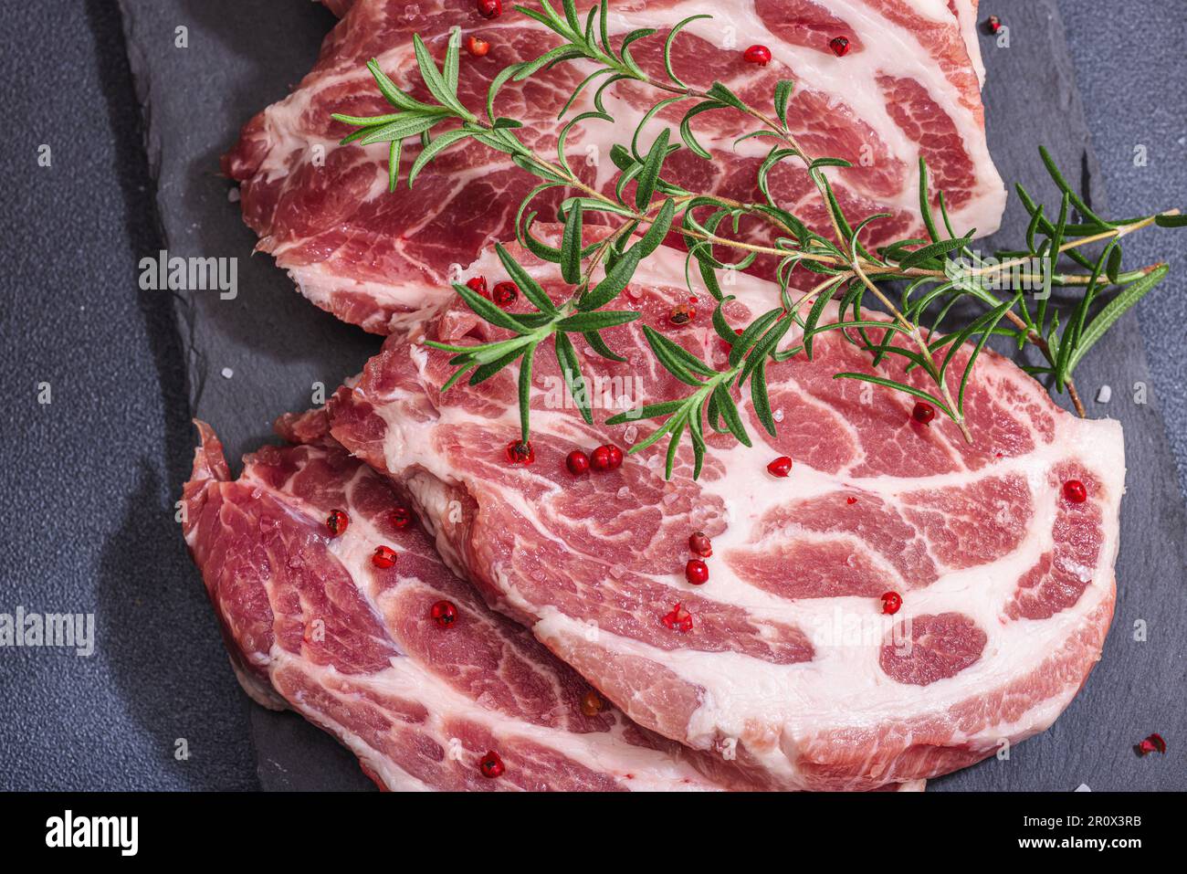 Rohes Schweinenackfleisch. Hacksteak, roter Pfefferkörner, Knoblauchzehen, Meersalz und frischer Rosmarin. Trendiger Standfuß, schwarzer Hintergrund, flach liegend, Draufsicht Stockfoto