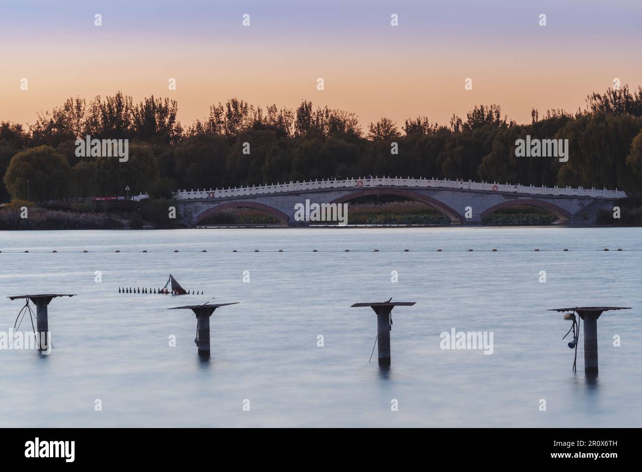 Ein ruhiger Sonnenuntergang beleuchtet einen malerischen Park mit einer bogenförmigen Brücke und Pfosten, die sich in das ruhige Wasser erstrecken Stockfoto