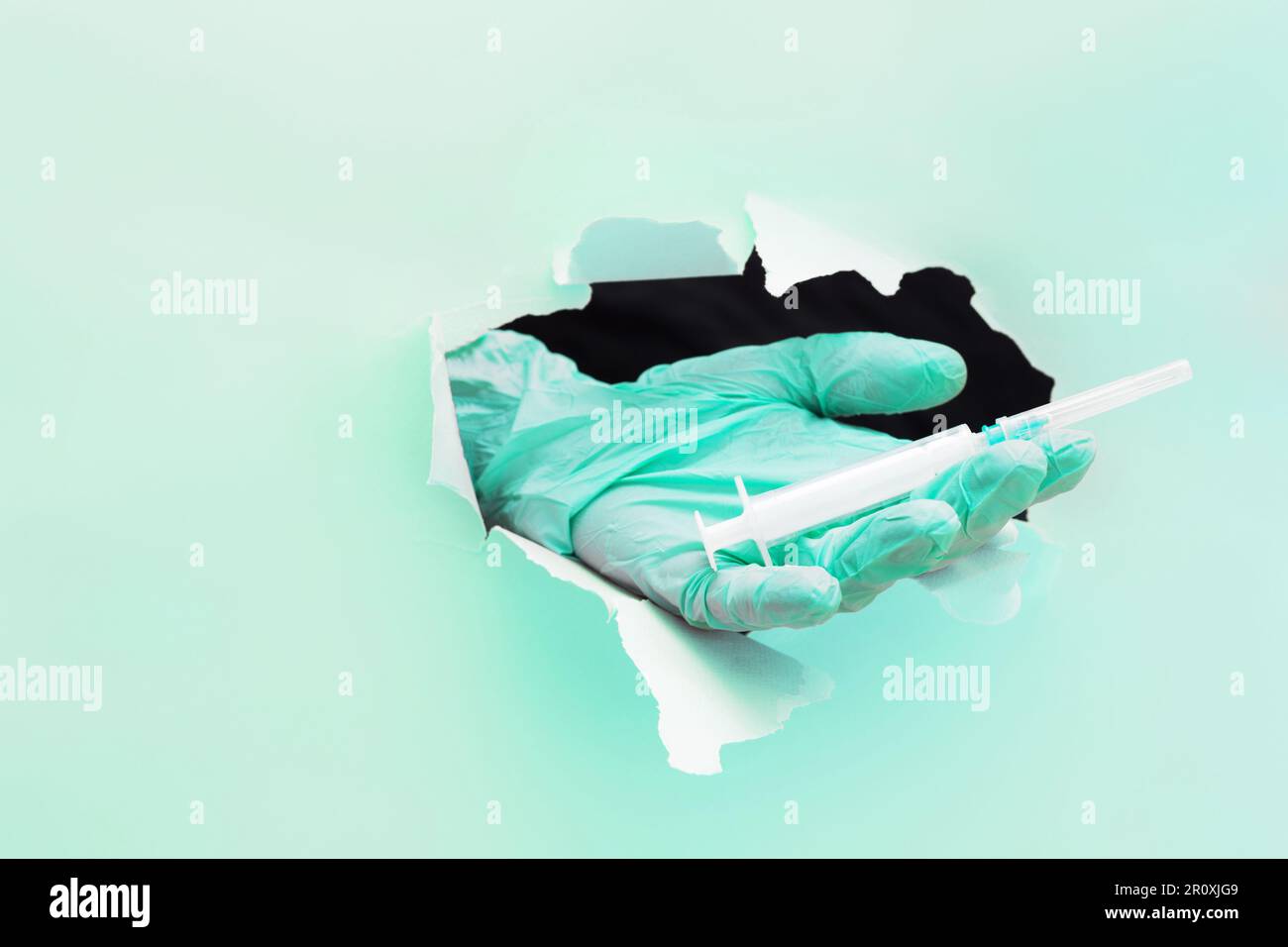 Die Hand eines Arztes hält eine Spritze mit einem Impfstoff durch ein Loch in zerrissenem Papier auf grünem Hintergrund Stockfoto