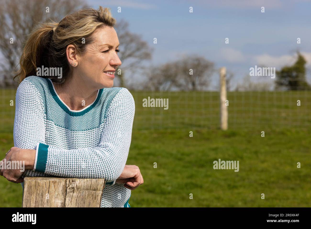 Attraktive, durchdachte Frau mittleren Alters, die sich auf einem Holzzaunpfahl auf dem Land oder in ihrem Garten ruht Stockfoto