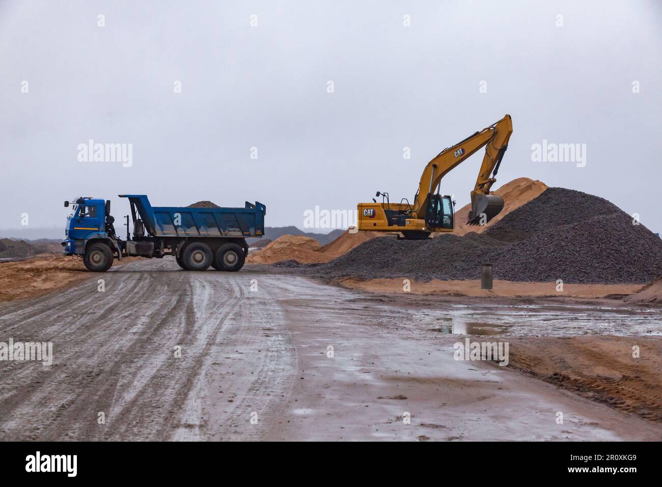 USt-Luga, Oblast Leningrad, Russland - 16. November 2021: Bagger Caterpillar lädt Kies in Muldenkipper. Nasse Fahrbahn im Vordergrund. Stockfoto