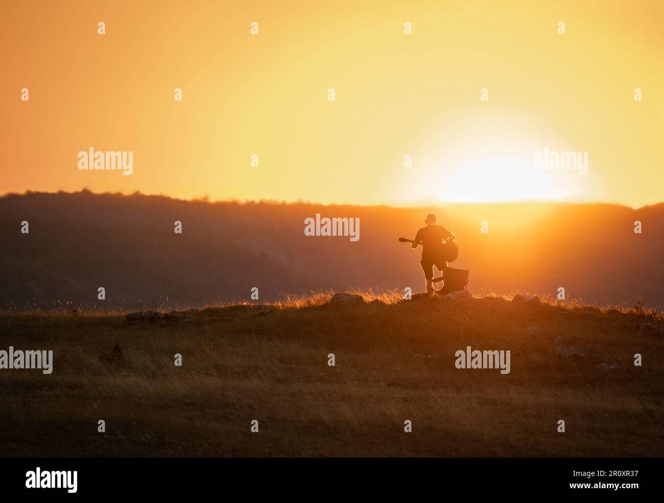 Silhouette eines Mannes, der bei Sonnenuntergang Gitarre spielt. Hügel im Hintergrund. Orangefarbener Himmel. Stockfoto
