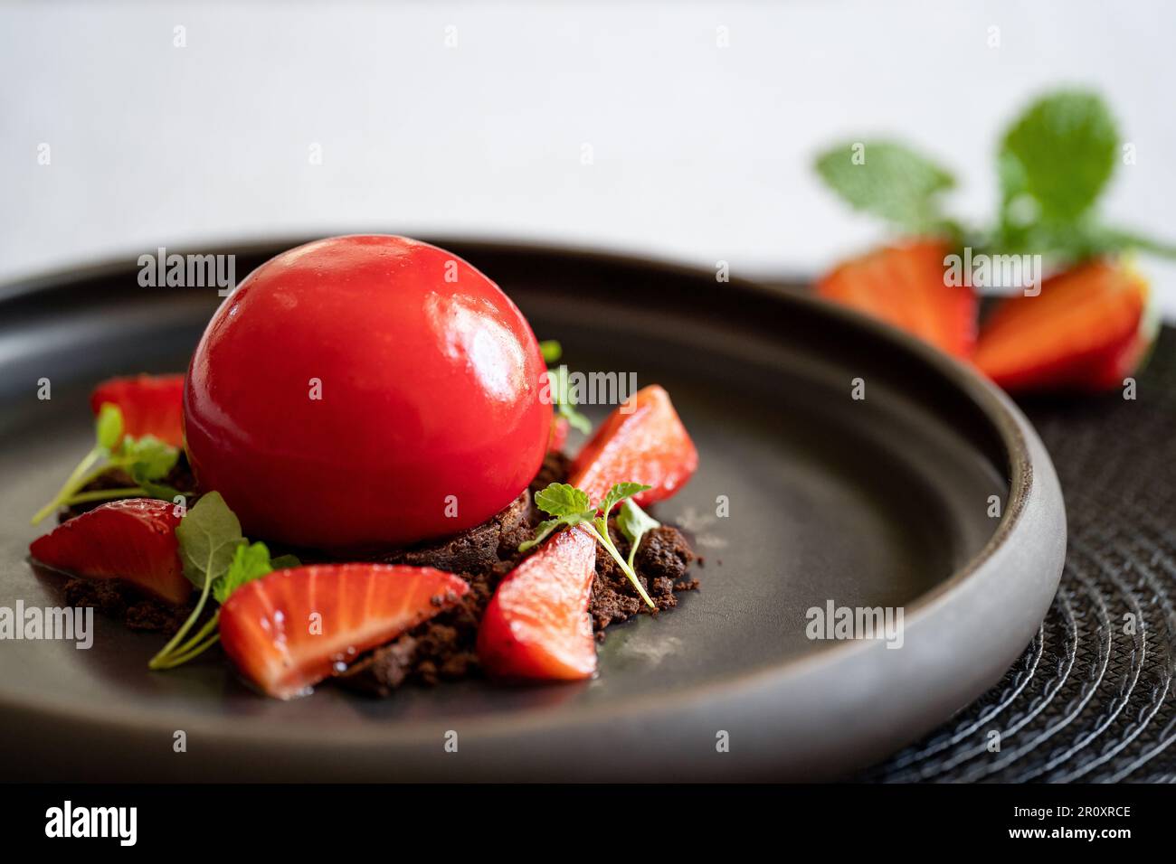 Selektivfokus eines kugelförmigen Bruchkuchens mit Erdbeeren und Brownie. Dessert mit glatten Oberflächen und Spiegelglasur. Rotes Dessert auf dem schwarzen Teller. Stockfoto