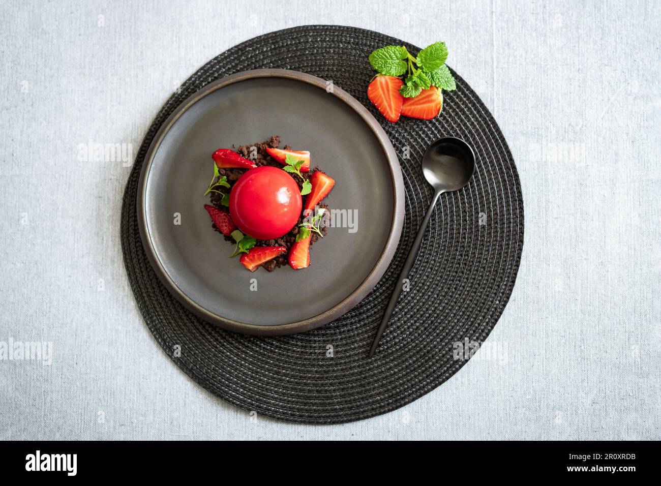 Draufsicht auf einen kugelförmigen Quark mit Erdbeeren und Brownie Crumbs. Rotes Dessert mit glatten Oberflächen und Spiegelglasur. Schwarzer Teller und Löffel. Stockfoto