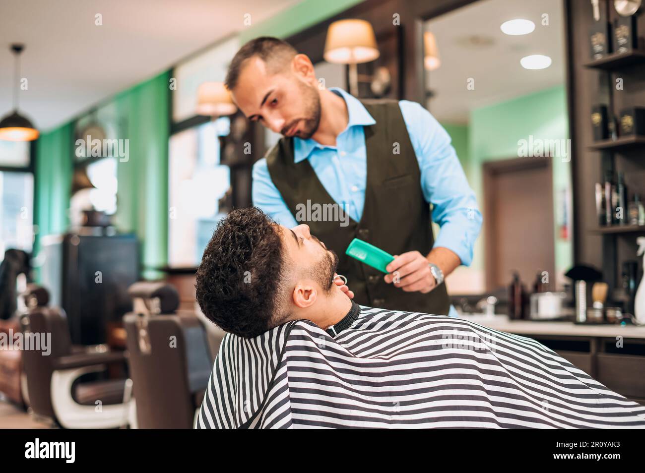 Männlicher Barbier mit Kammschnitt mit Schere des Kunden in gestreiftem Umhang während des Haarschnitts in einem modernen Friseursalon Stockfoto