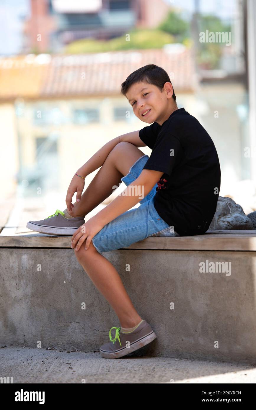 Ein Junge in Shorts und schwarzes T-Shirt, der auf einer Steinbank sitzt und in die Kamera schaut. Stockfoto