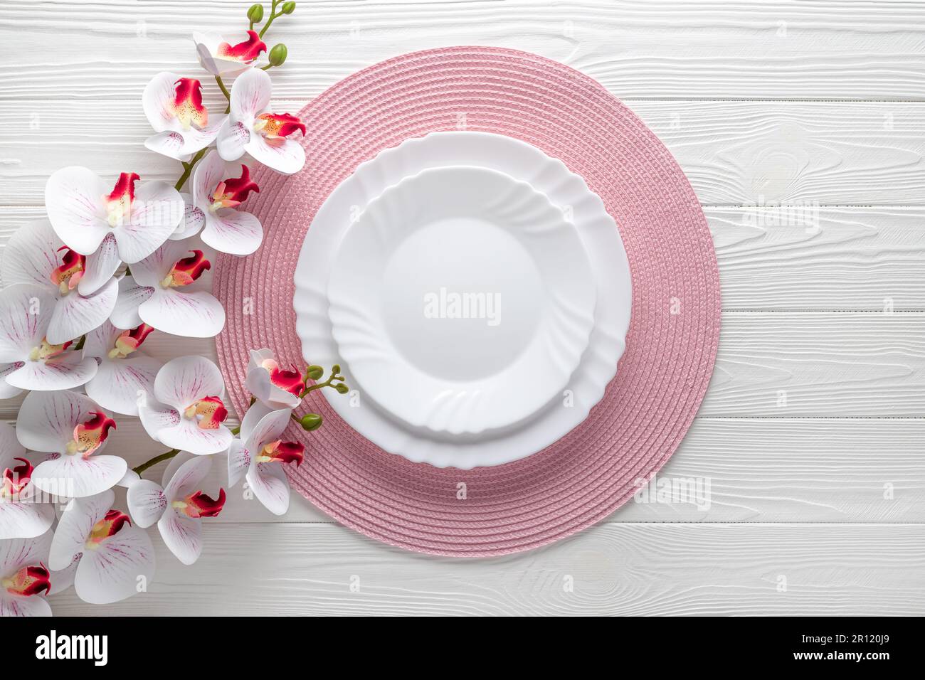 Festliches Ambiente mit pinkfarbenen Servietten und Orchideenblumen. Leere Teller auf weißem Holztisch. Draufsicht. Festliches Abendessen. Karte oder Menüvorlage, flach d Stockfoto