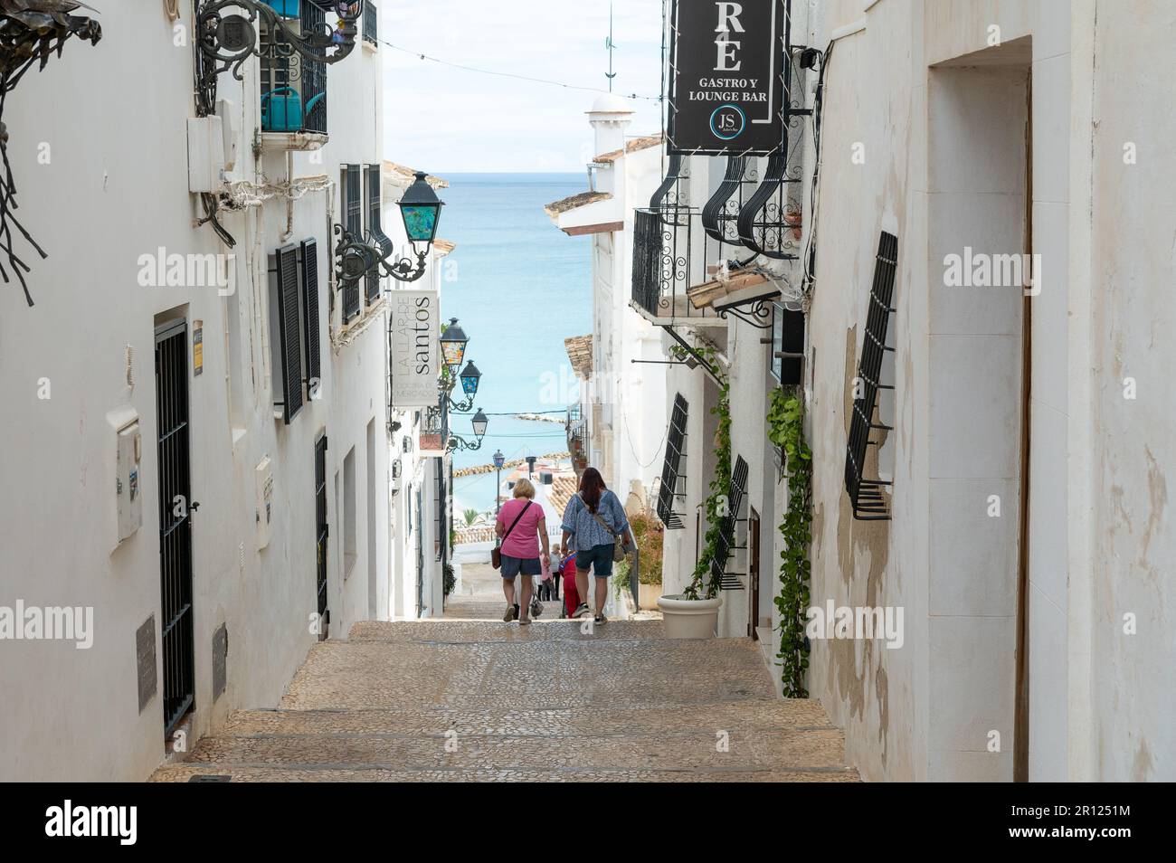 Die Leute gehen einen steilen Pfad zwischen zwei Gebäuden mit Meer im Hintergrund in Altea, Spanien. Stockfoto