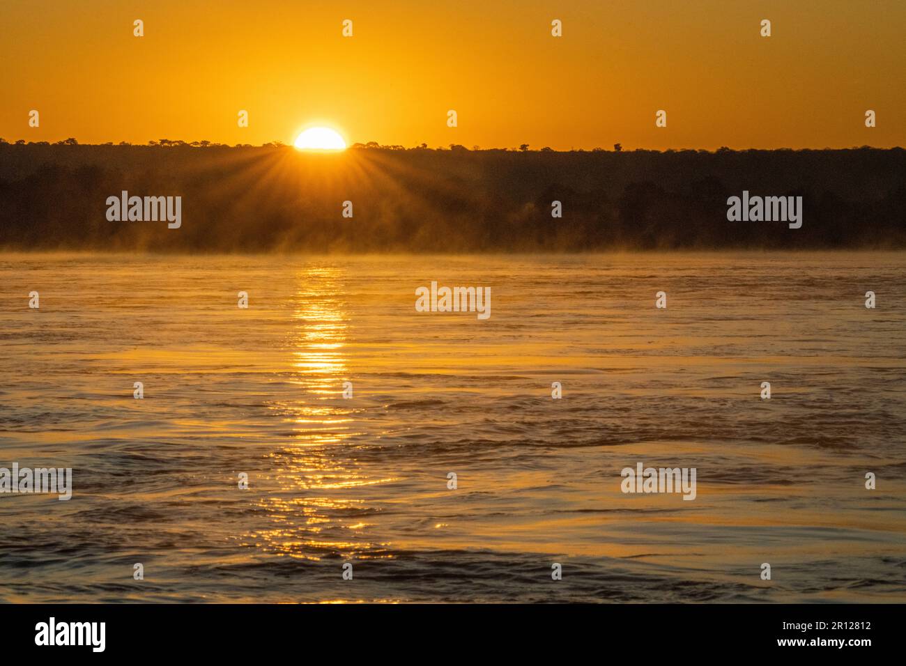 Sonnenaufgang über dem Sambesi-Fluss, die Wasseroberfläche leuchtet in leuchtenden orangefarbenen Farben auf, Nebel steigt aus dem Wasser auf. Sambesi, Simbabwe, Afrika Stockfoto