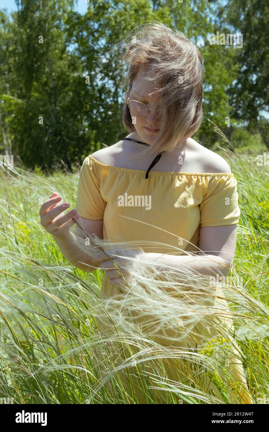 Asiatisches Mädchen mit Strohhut und gelbem Kleid inmitten des flauschigen Grases auf dem Feld. Sommerzeit. Dorfurlaub. Zeit zu reisen Stockfoto