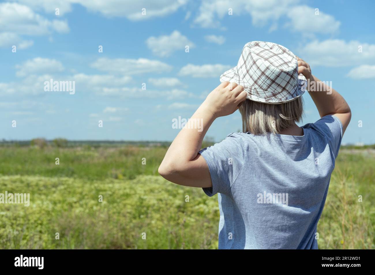 Eine Frau mit Hut und einem blauen T-Shirt auf einem Feld, blauer Himmel mit Wolken, Rückansicht. Sommerzeit. Dorfurlaub. Zeit zu reisen Stockfoto