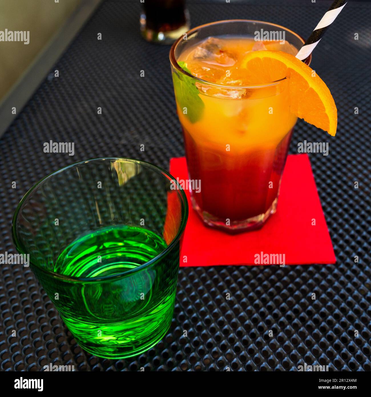 Saftiger orange-roter Cocktail mit gestreiftem Stroh, Wasser in grünem Glas, rote Serviette auf schwarzem Tisch. Stockfoto