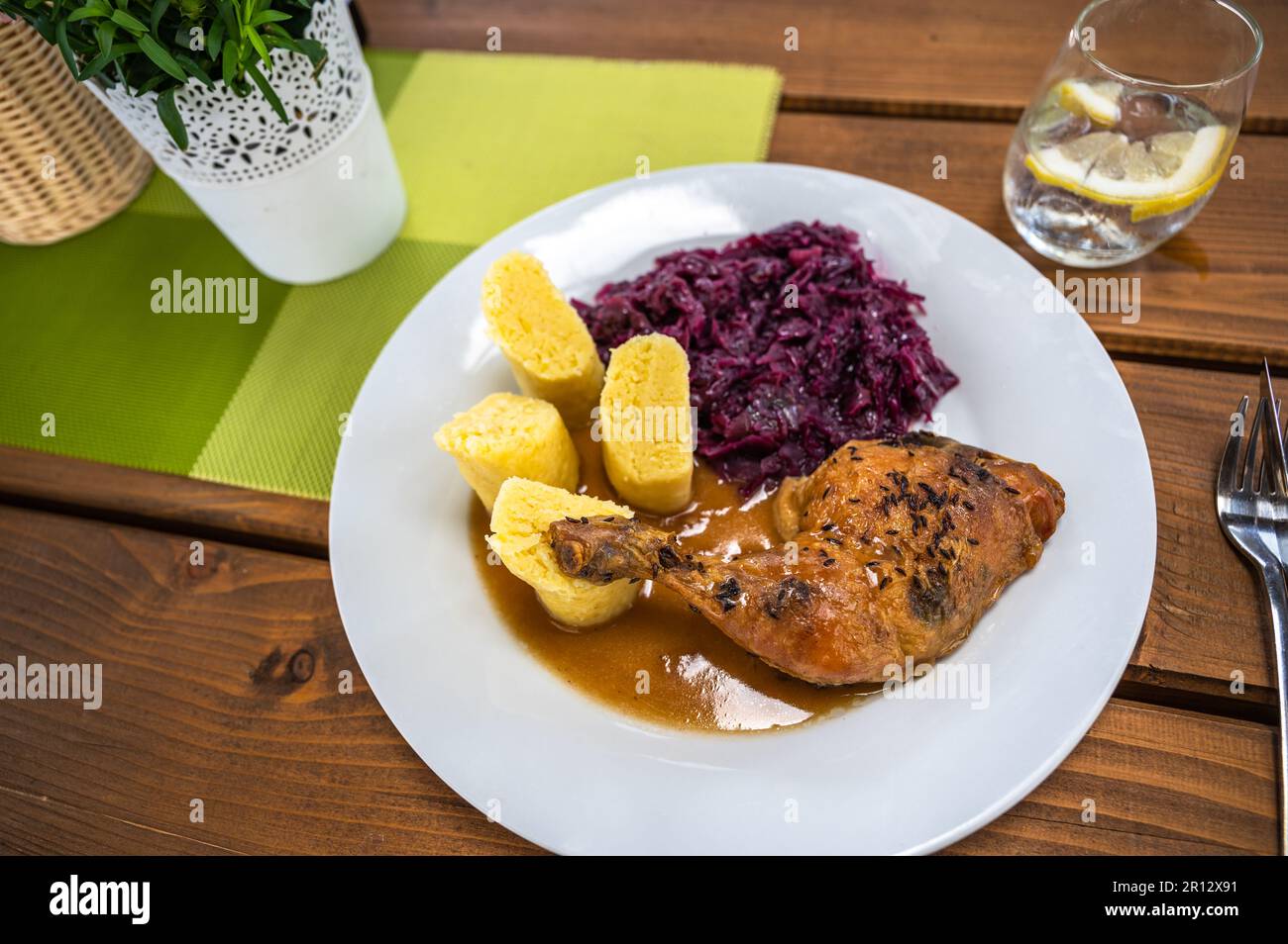 Gebackene Hühnerschenkel, gedünsteter Rotkohl und flauschige Kartoffelklöße, weißer Teller, Glas Getränk, Topf mit Blume, Tischdecke auf dem Tisch, Nahaufnahme. Stockfoto