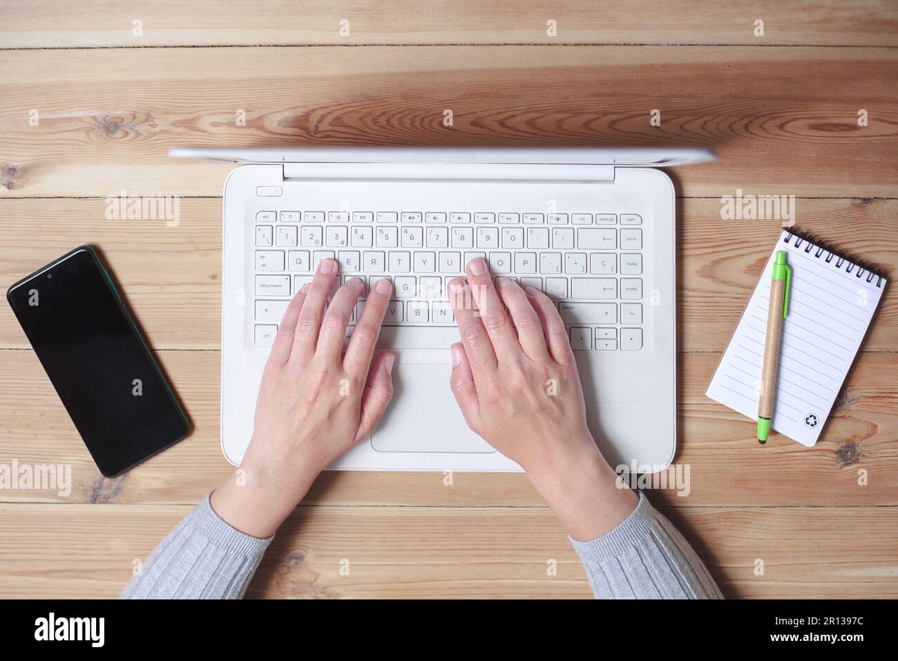 Draufsicht der Hände einer Frau, die auf einem Laptop tippen. Daneben befinden sich ihr Smartphone und ein Notizblock. Stockfoto