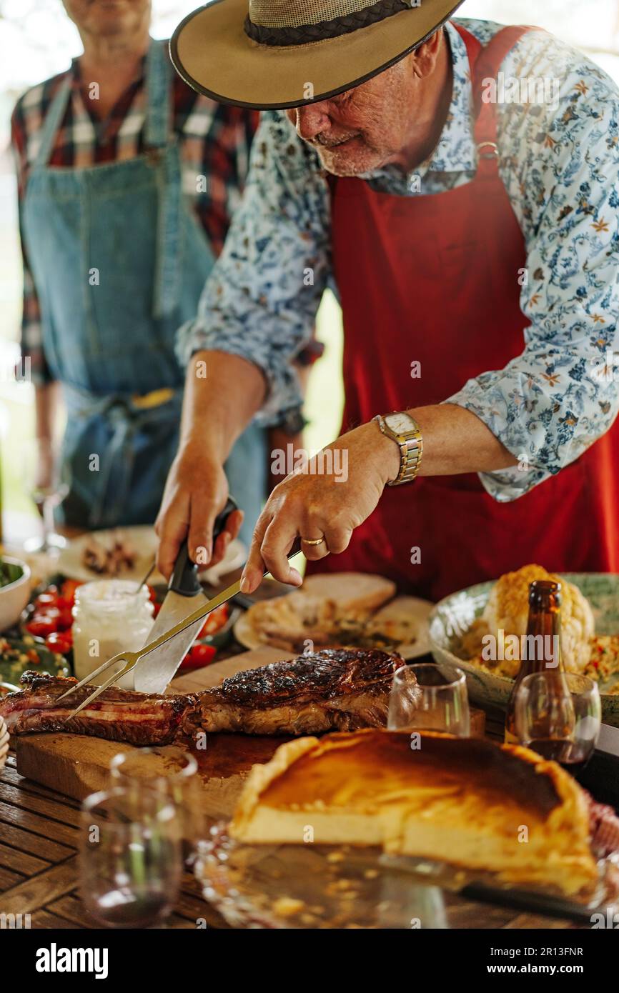 Ein 65-jähriger Mann mit Strohhut und Schürze schneidet gegrilltes Fleisch geschickt mit einem Barbecue-Messer und einer Gabel. Im Hintergrund, nicht wiedererkennbare Menschen Stockfoto