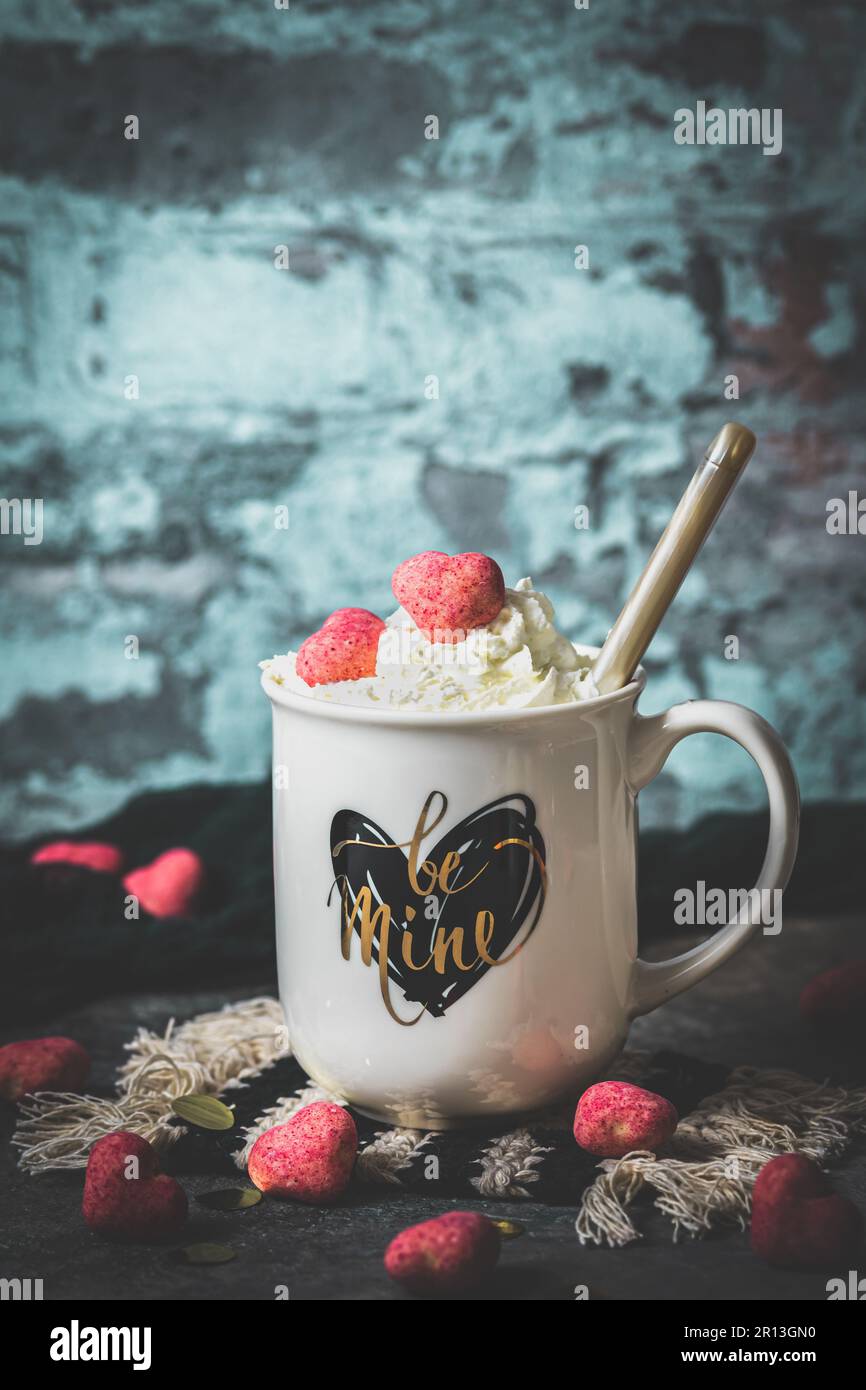 Becher mit Herzmuster und Text sei meins, gefüllt mit Kaffee oder Kakao mit Schlagsahne und rosa Schokoladenherzen, rustikaler Hintergrund, Kopierbereich Stockfoto