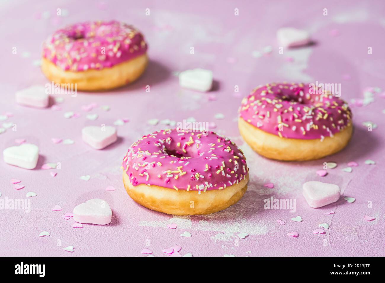 Donuts, Berliner, Krapfen mit rosafarbener Zuckerzusatz auf pinkfarbenem strukturiertem Hintergrund, dekoriert mit pinkfarbenem und weißem Zucker und Papierherzen Stockfoto