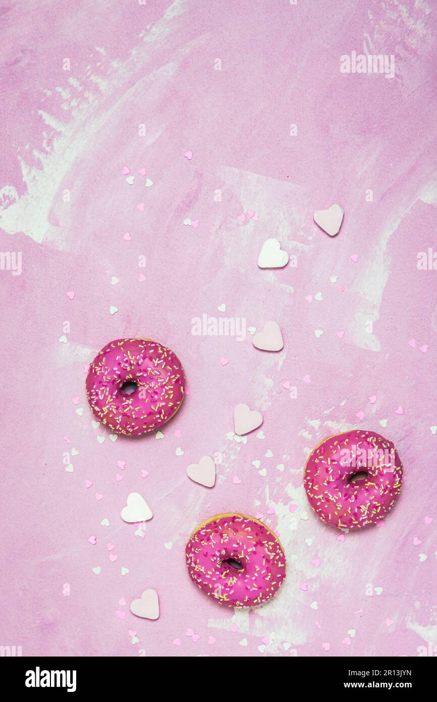 Donuts, Berliner, Krapfen mit rosafarbener Zuckerglasur auf pinkfarbenem strukturiertem Hintergrund, dekoriert mit pinkfarbenem und weißem Zucker und Papierherzen, Draufsicht mit Kopie Stockfoto