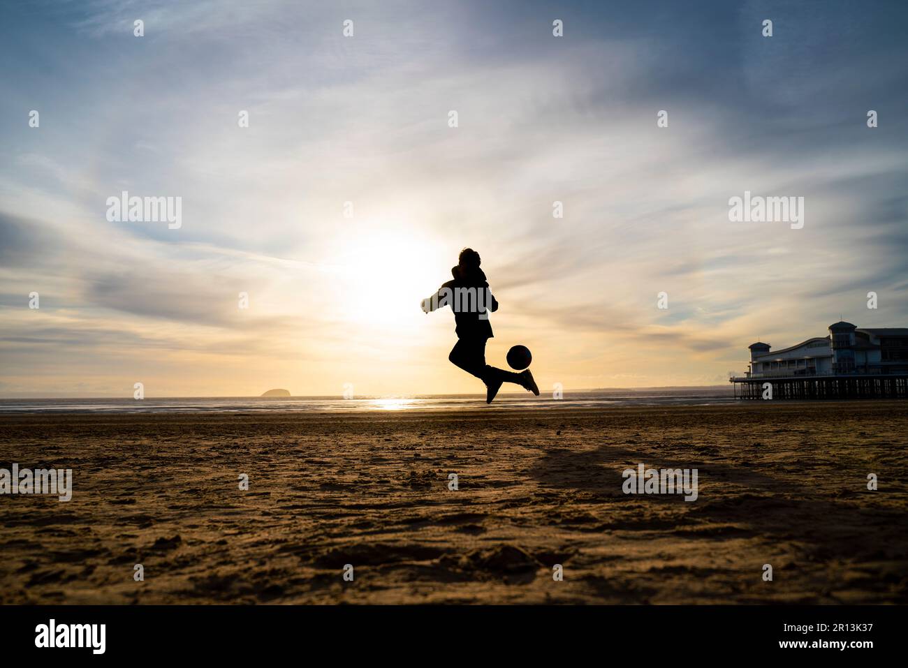 Silhouette eines kleinen Jungen, der bei Sonnenuntergang am Strand in Weston-super-Mare, Somerset, Großbritannien, Fußball spielt. Stockfoto