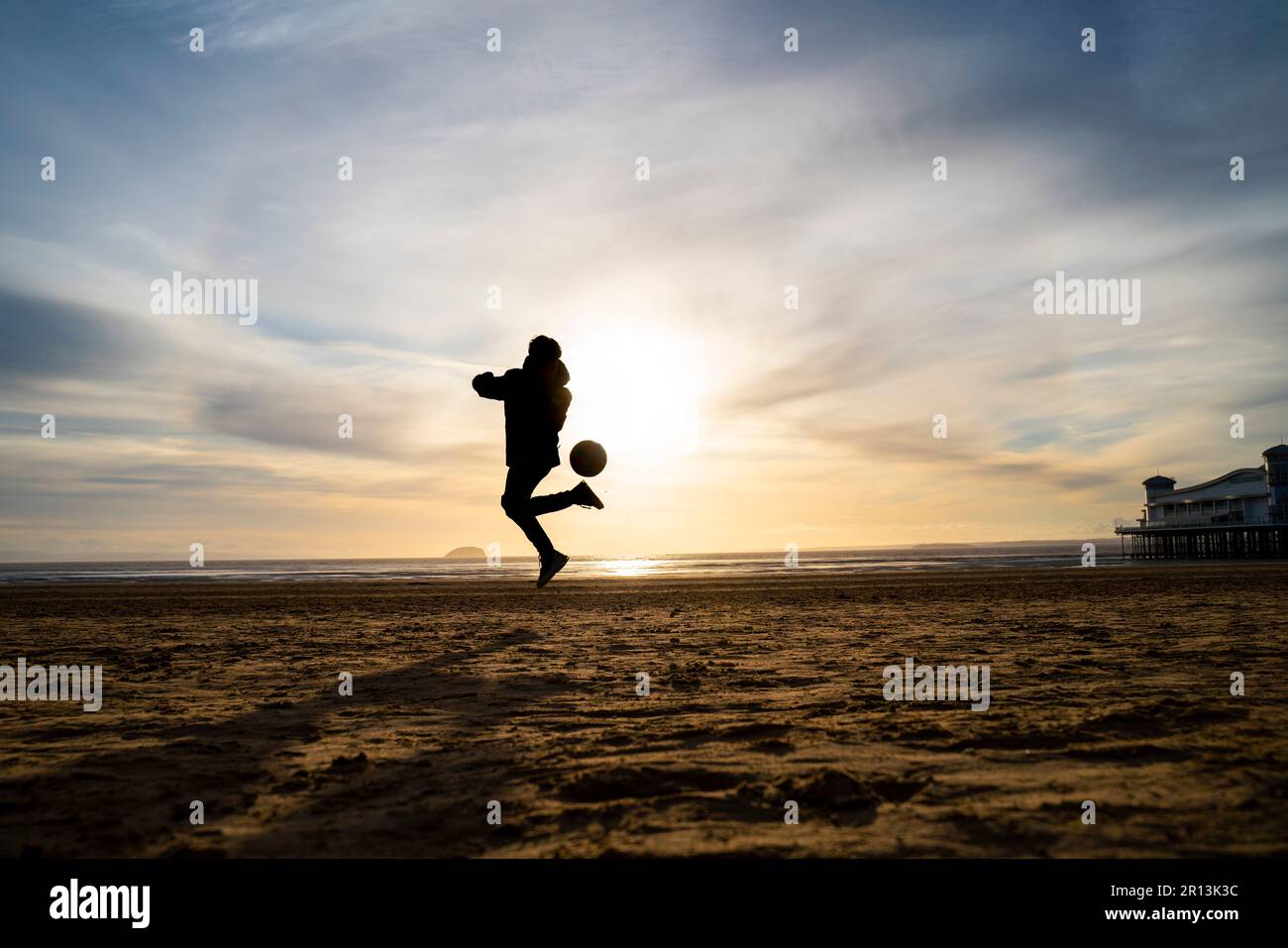 Silhouette eines kleinen Jungen, der bei Sonnenuntergang am Strand in Weston-super-Mare, Somerset, Großbritannien, Fußball spielt. Stockfoto