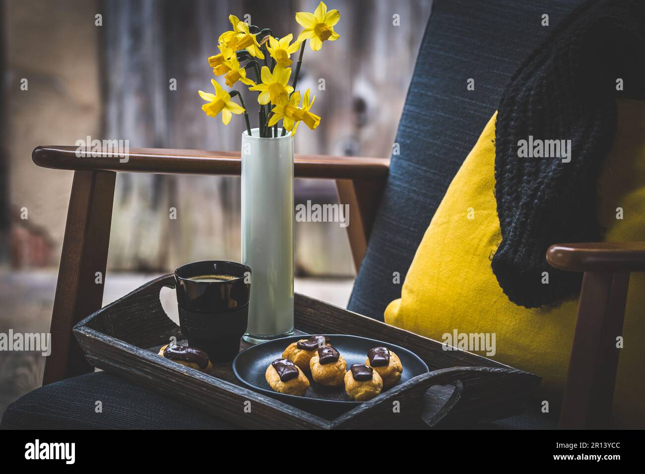 Holztablett mit Mini-Eclairs und Kaffee auf einem Sessel vor dem Fenster, Kaffeepause, vertikal Stockfoto