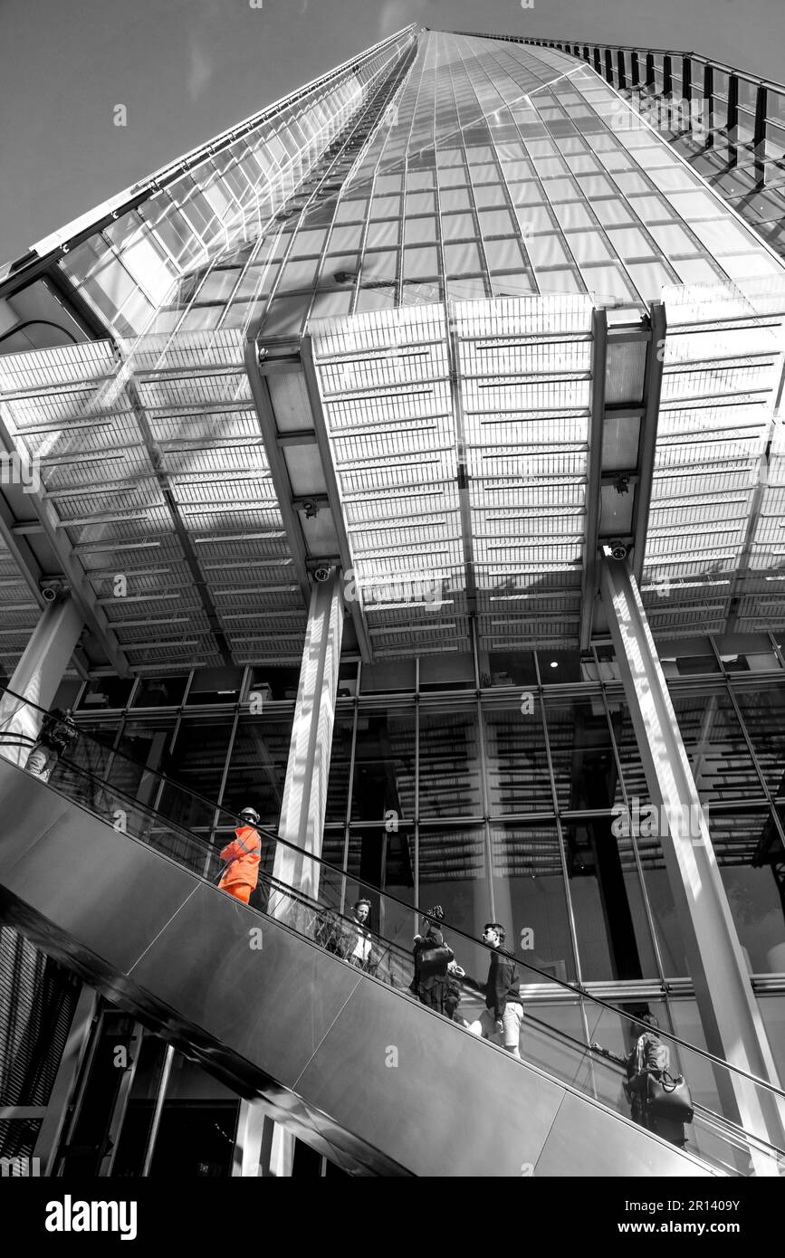Leute, die die Rolltreppe nach The Shard in London, England, hochfahren. Balck und Weiß mit farbigem Burst auf gut sichtbarer Jacke eines Werkers. Stockfoto
