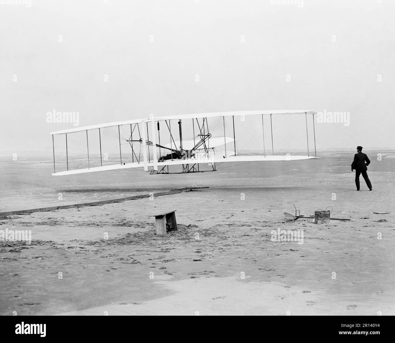 Erster Flug, 120 Fuß in 12 Sekunden; Kitty Hawk, North Carolina. Das Foto zeigt Orville Wright an den Bedienelementen der Flugmaschine, die in Bauchlage auf dem unteren Flügel liegt, mit Hüften in der Wiege, die den Flügelmechanismus betätigt. Wilbur Wright, der zur Waage gerannt ist, hat gerade seinen Griff an der vorderen Stütze des rechten Flügels gelöst. Hinter der Maschine sind die Startschiene, die Flügelstütze, ein Spulenkasten und andere für die Flugvorbereitung erforderliche Teile sichtbar. Foto von John T. Daniels Stockfoto
