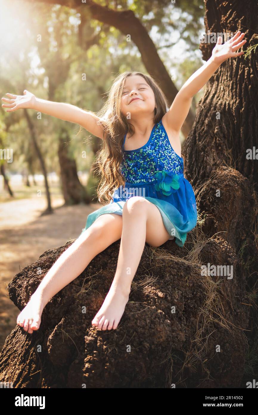 Wunderschönes glückliches Mädchen, das im Park mit blauem Kleid ohne Schuhe  spielt, auf dem Baum sitzt und vor Glück schreit, während die Familie  Kinder genießt Stockfotografie - Alamy