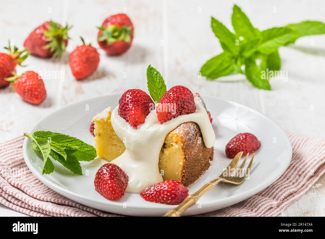 Ein Stück hausgemachter Käsekuchen mit Schlagsahne und frischen Erdbeeren auf weißem Holzhintergrund, dekoriert mit frischen Minzblättern Stockfoto