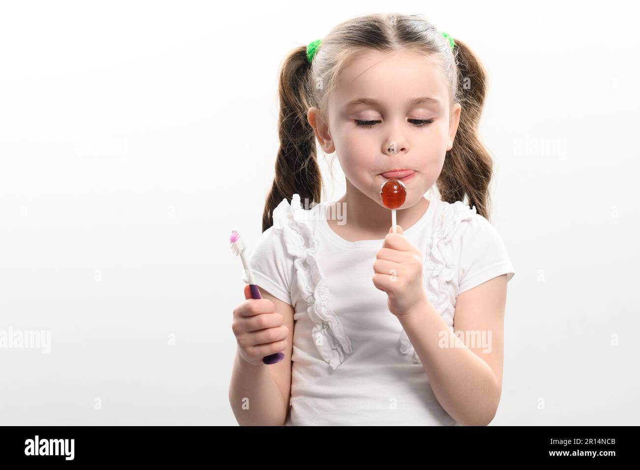 Das kleine Mädchen leckt einen Lutscher und hält eine Zahnbürste auf weißem Hintergrund, Kopierbereich. Stockfoto