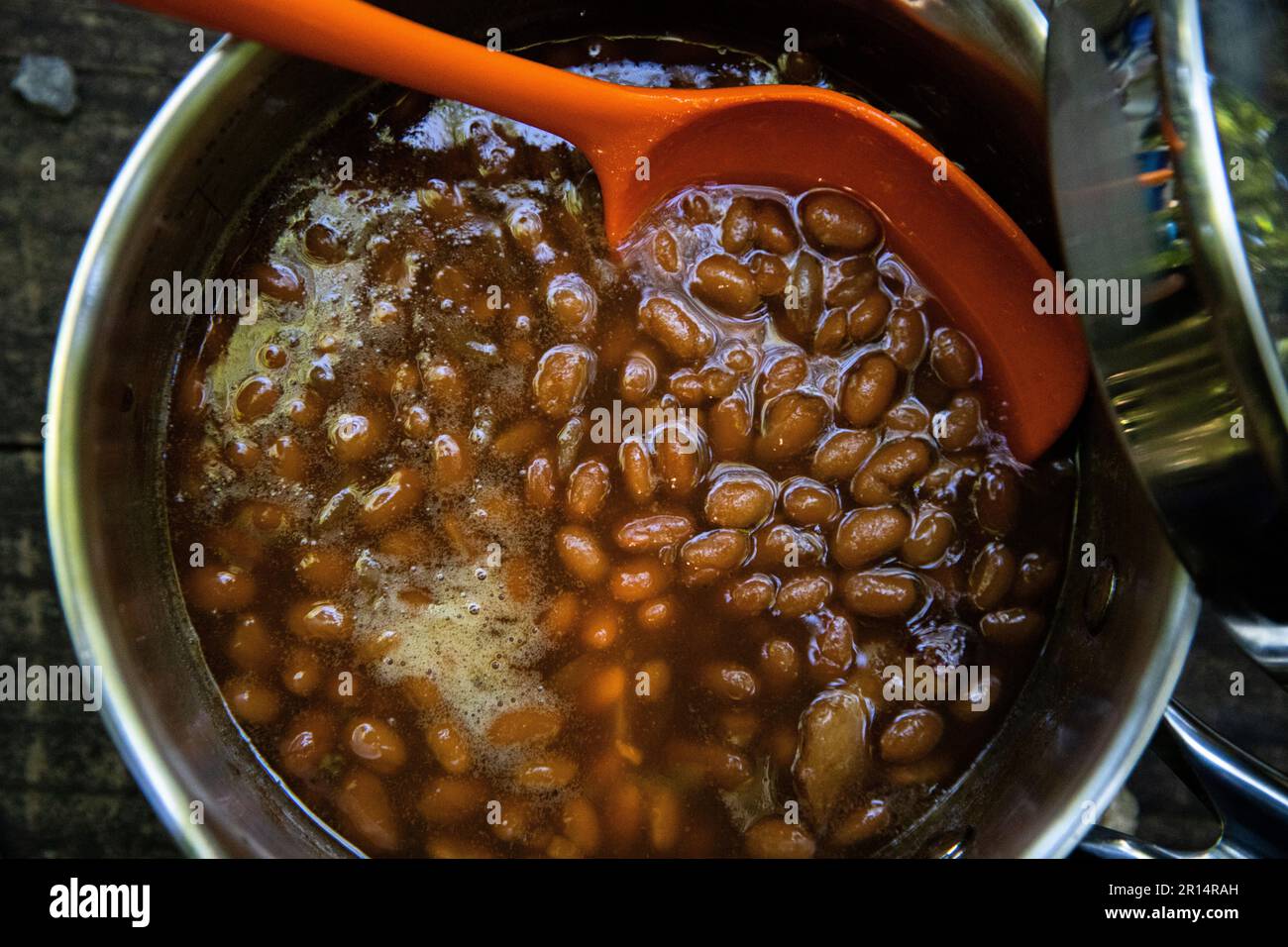 Nahaufnahme von gebackenen Bohnen in einem Gericht auf dem Tisch mit einem orangefarbenen Servierlöffel, perfektes Sommergericht beim Kochen oder Treffen Stockfoto