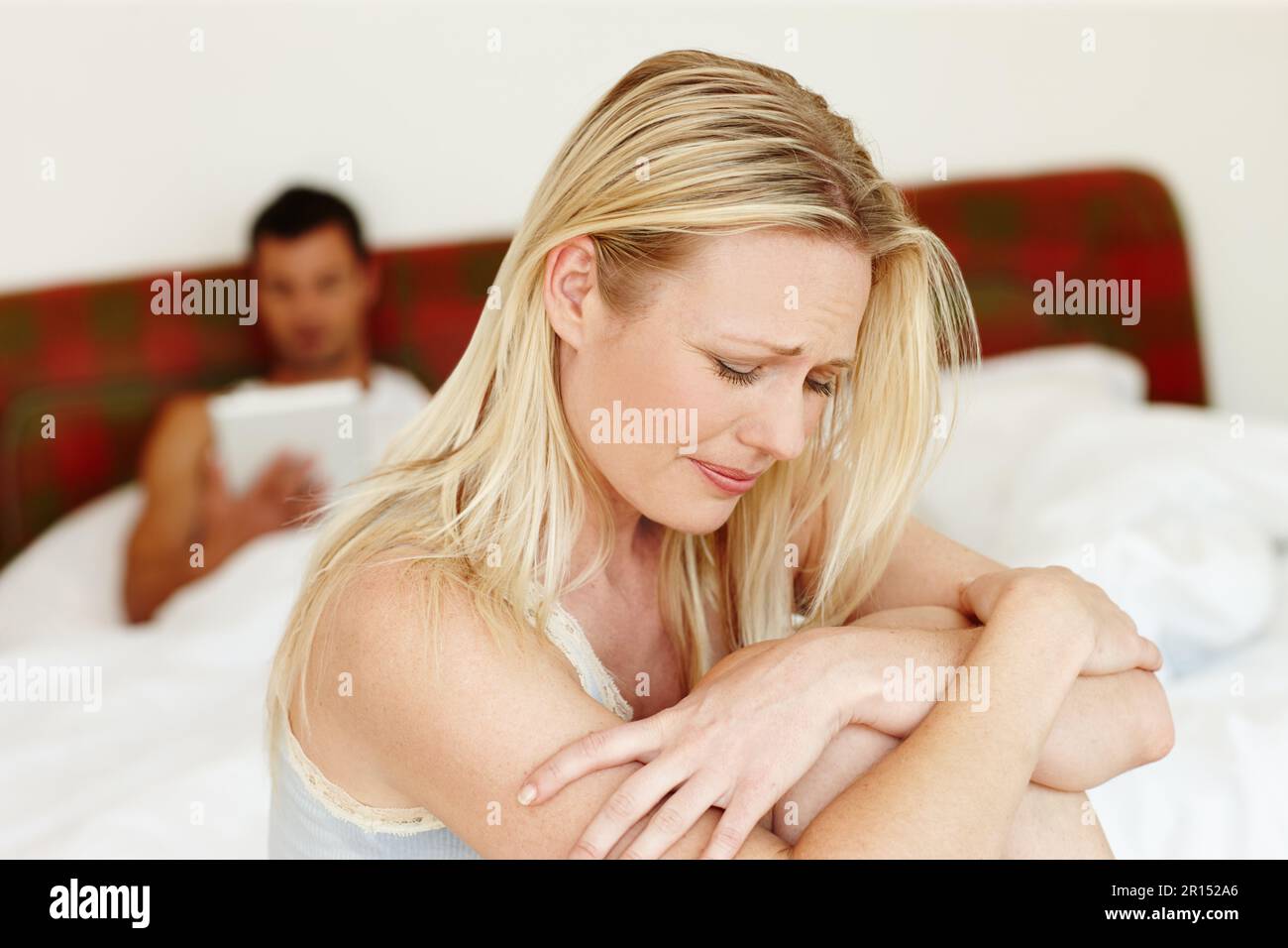 Die Risse zeigen sich in unserer Beziehung. Eine aufgebrachte Frau, die auf dem Rand ihres Bettes sitzt, während ihr Mann im Hintergrund liegt. Stockfoto