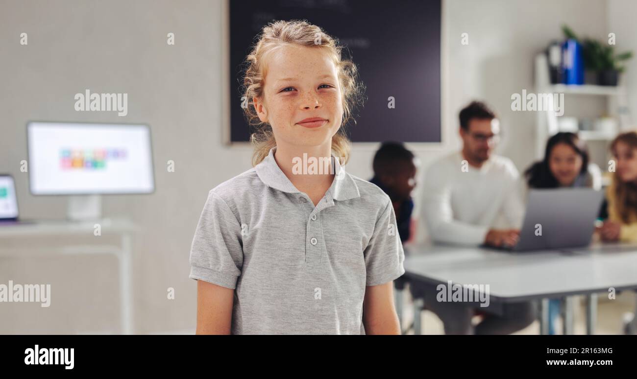 Männlicher Schüler, der in einem Klassenzimmer in die Kamera schaut, während sein Lehrer mit anderen Schülern im Hintergrund arbeitet. Junger Junge, der in einer Umgebung zur Schule geht Stockfoto