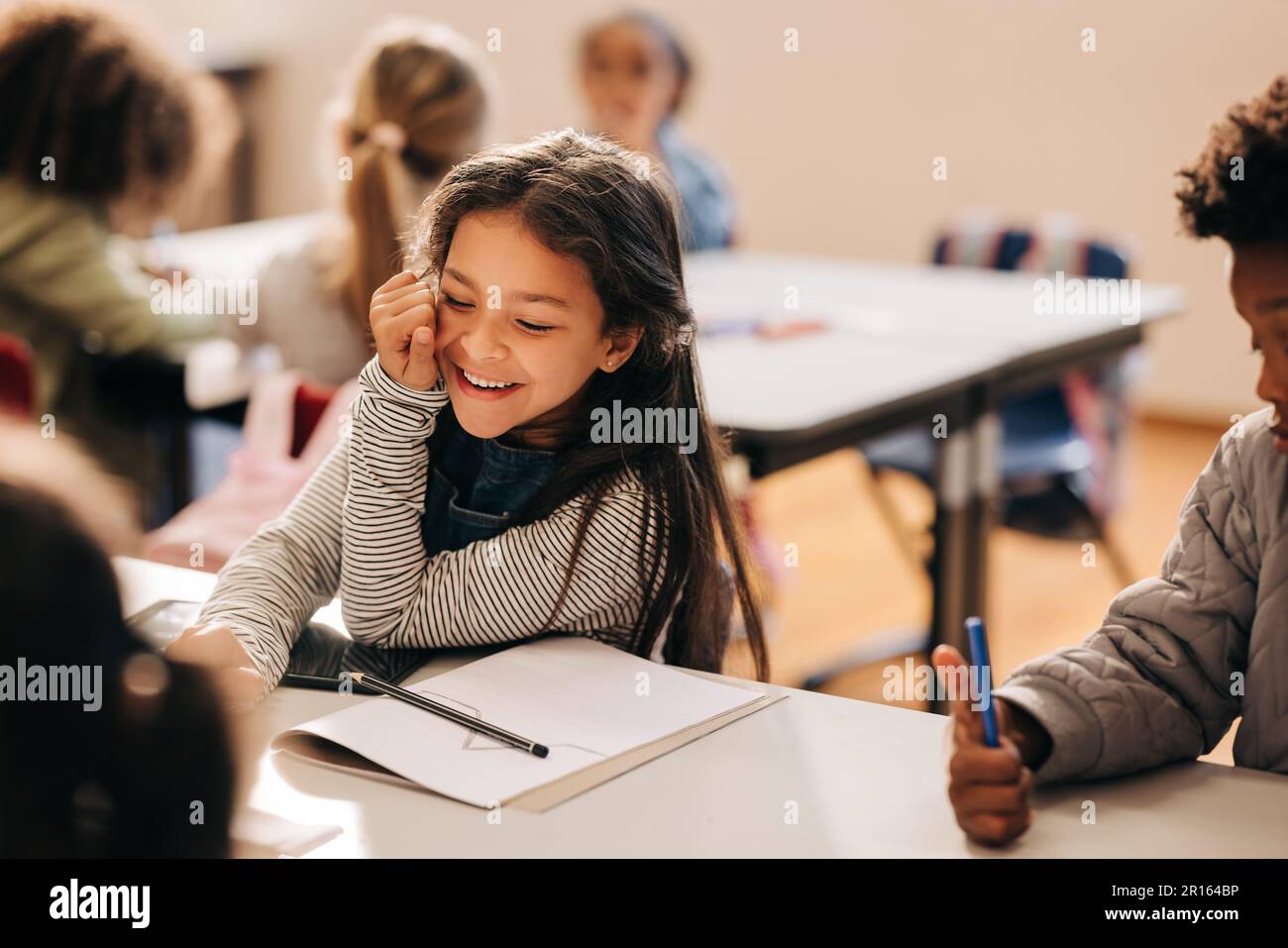 Ein kleines Mädchen lacht glücklich in einer Grundschule, sie sitzt mit ihren Klassenkameraden an einem Tisch. Grundschüler genießen Kunst und Stockfoto