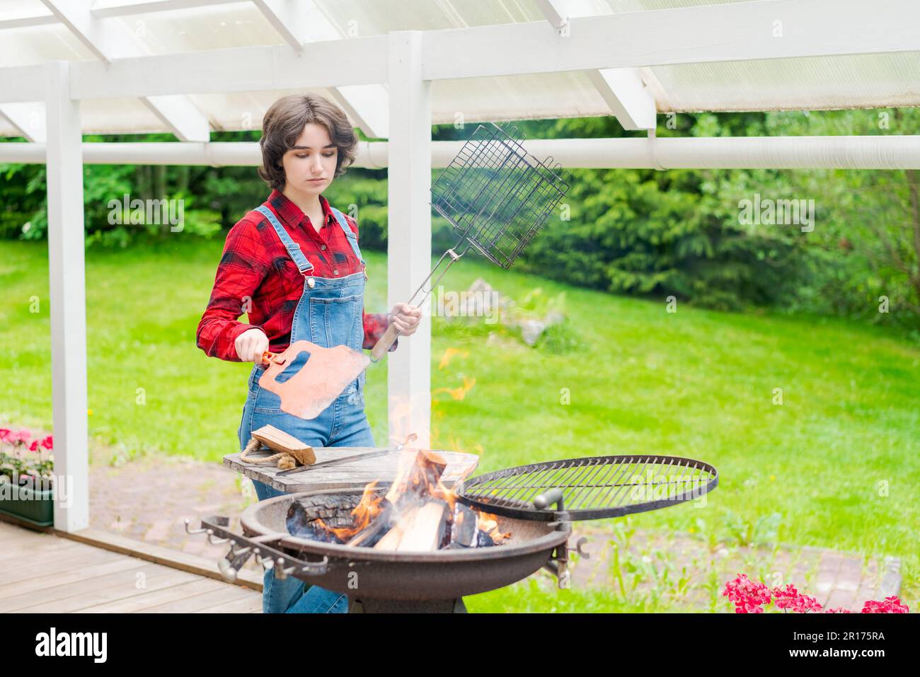 Grillparty im Garten mit einer jungen Frau in Jeansanzug und rotem Karohemd in einem Landhaus auf der Terrasse, das Fleischspieße zubereitete Stockfoto