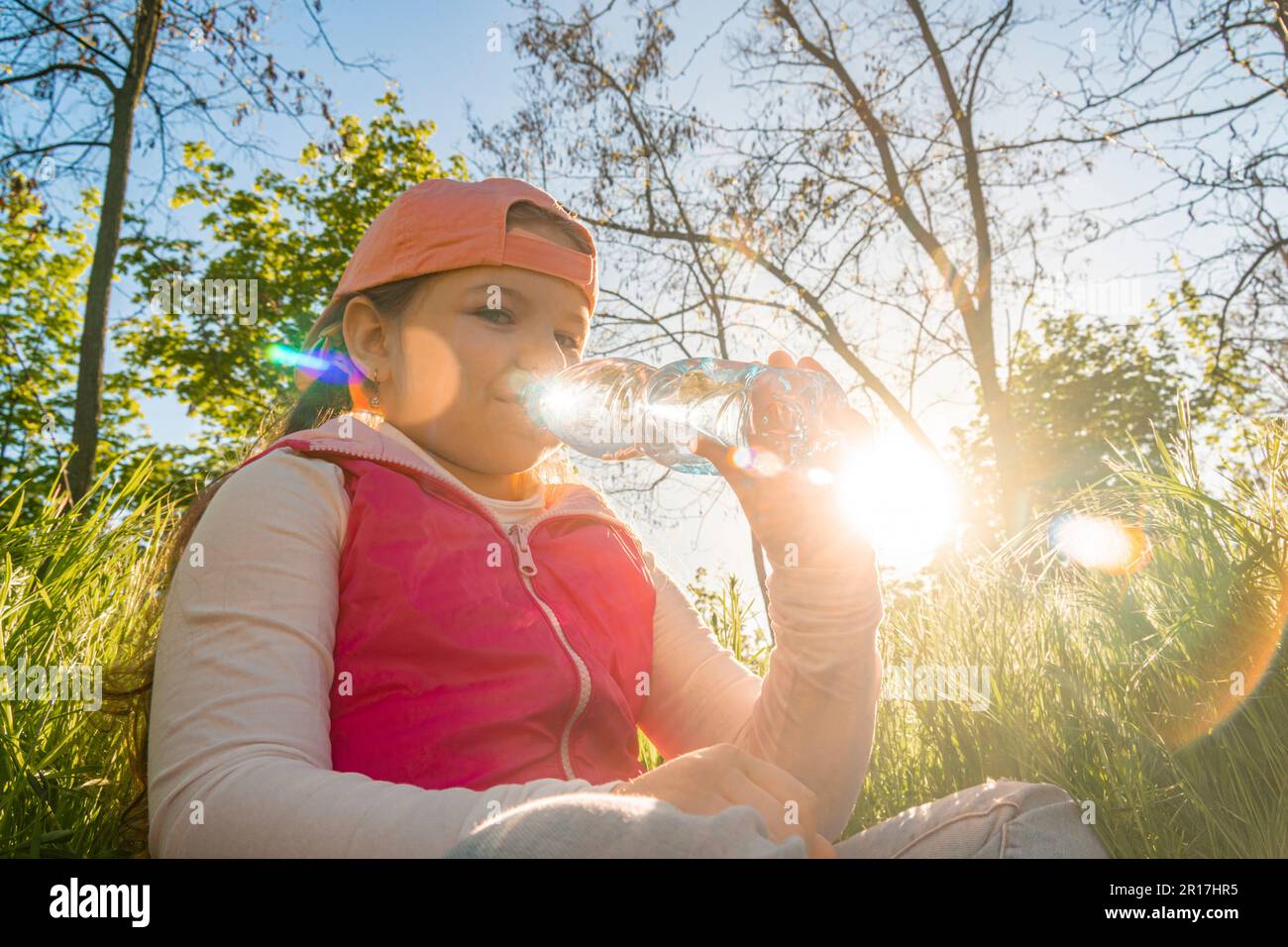 Ein Mädchen sitzt im dicken grünen Gras und trinkt Wasser aus einer Flasche vor dem Hintergrund des Sonnenuntergangs. Machen Sie einen Spaziergang im Stadtpark Stockfoto