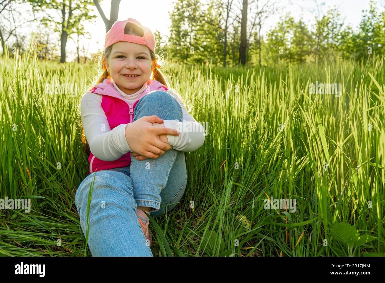 Ein fröhliches Mädchen sitzt auf dem dicken grünen Gras und hält ein Bein mit der Hand fest, vor dem Hintergrund des Sonnenuntergangs. Machen Sie einen Spaziergang im Stadtpark Stockfoto