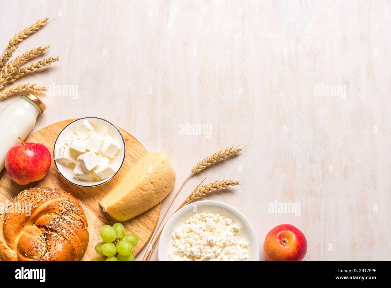 Happy Shavuot – religiöses jüdisches Urlaubskonzept mit Milchprodukten, Käse, traditionellem Brot, Milchflaschen, Weizen und Früchten auf weißem Holzrücken Stockfoto