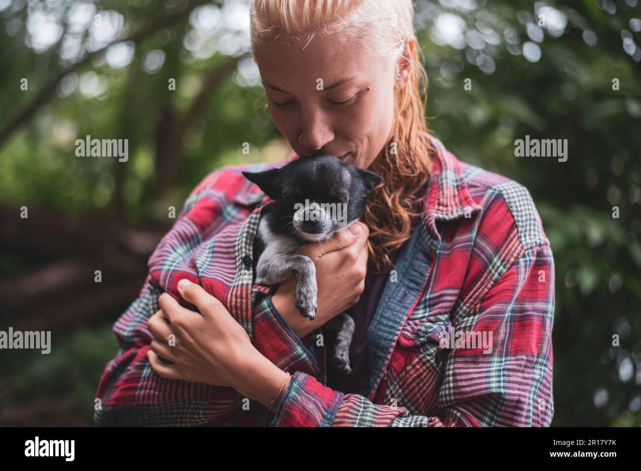 Eine blonde Frau, die einen kleinen süßen Hund im Grünen Wald hält und lächelt Stockfoto