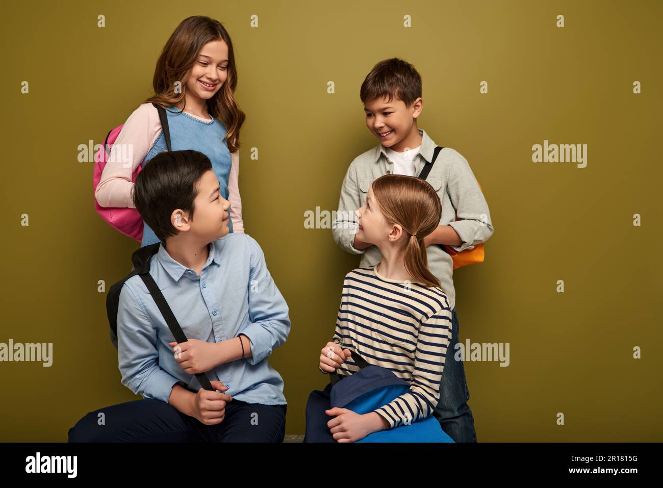 Positive multiethnische Schulkinder, die sich mit jungen Freunden unterhalten, während sie Rucksäcke in der Hand halten, sich anschauen und internationale Kinder da feiern Stockfoto