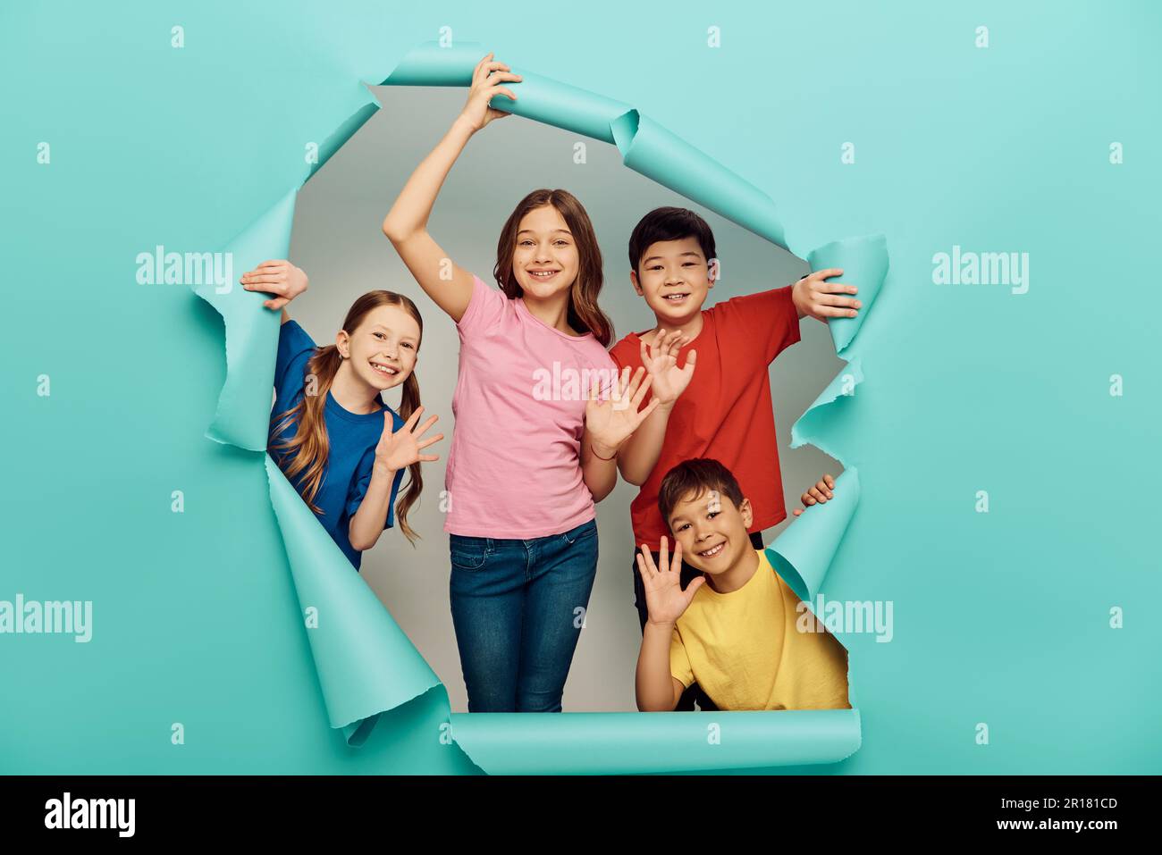 Lächelnde Kinder unterschiedlicher Rassen in lässiger Kleidung, die während des internationalen Kindertags mit den Händen vor der Kamera schwenken, hinter einem Loch in blauem Papierhintergrund Stockfoto