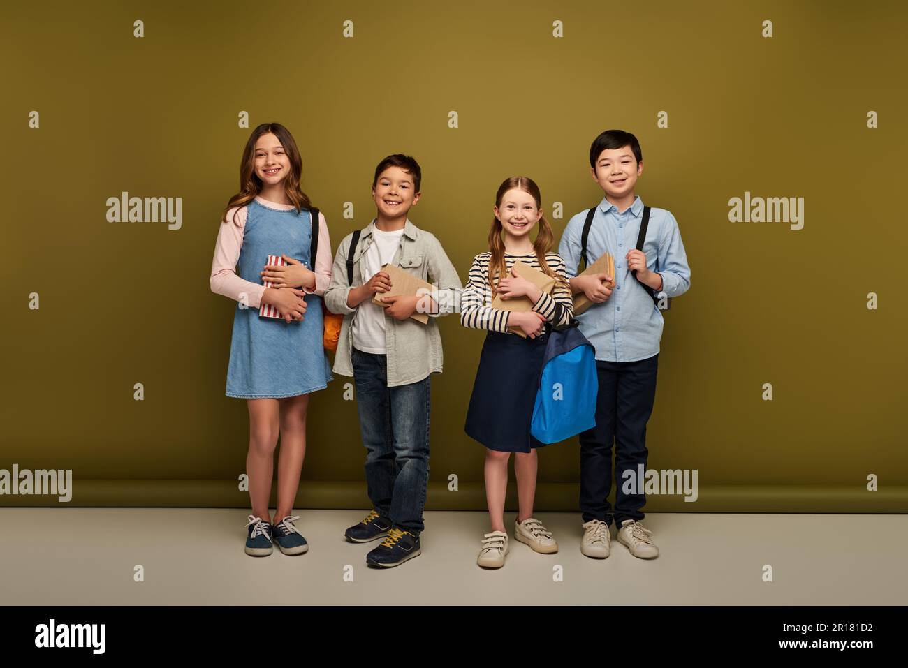 Die ganze Länge voller fröhlicher multiethnischer Kinder in legerer Kleidung mit Rucksäcken und Büchern auf Khaki-Hintergrund, Happy Children's Day Konzept Stockfoto