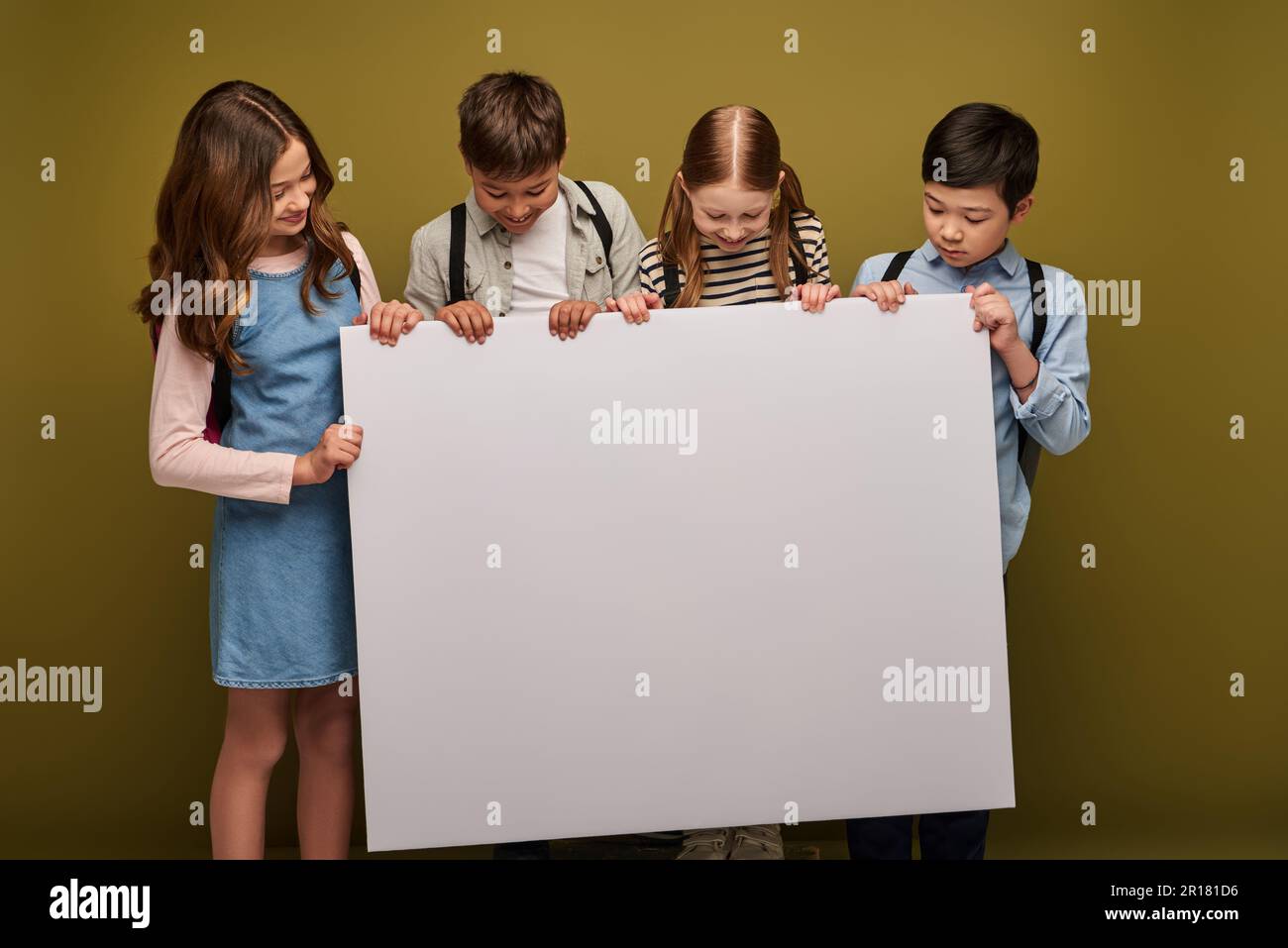 Positive multiethnische Kinder mit lächelnden Rucksäcken, während sie zusammenstanden und auf ein leeres Plakat auf Khaki-Hintergrund blickten, Happy children's d Stockfoto