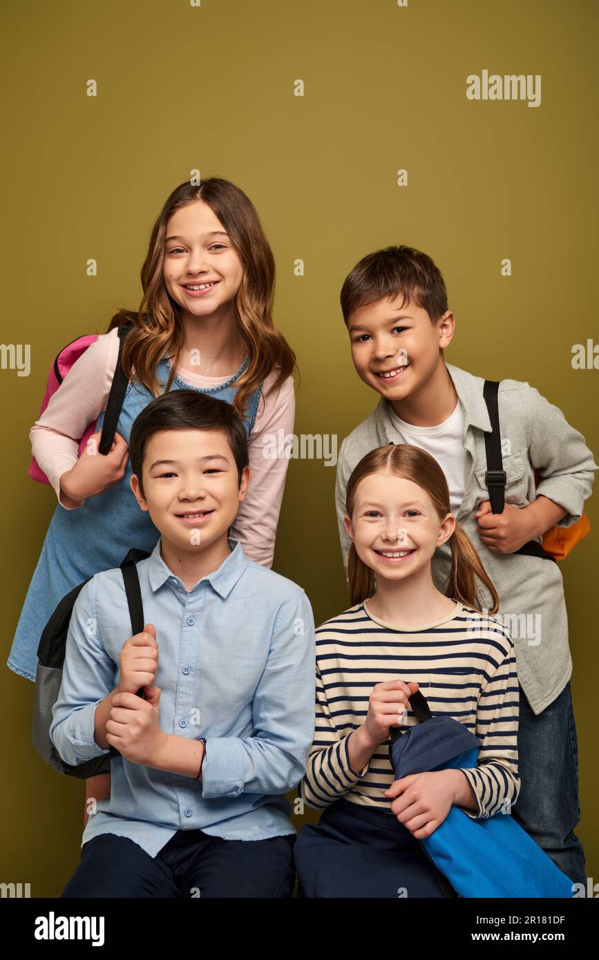 Lächeln multiethnischer Kinder in lässiger Kleidung, mit Rucksäcken und Blick in die Kamera während der Kinderschutzfeier auf Khaki-Hintergrund Stockfoto