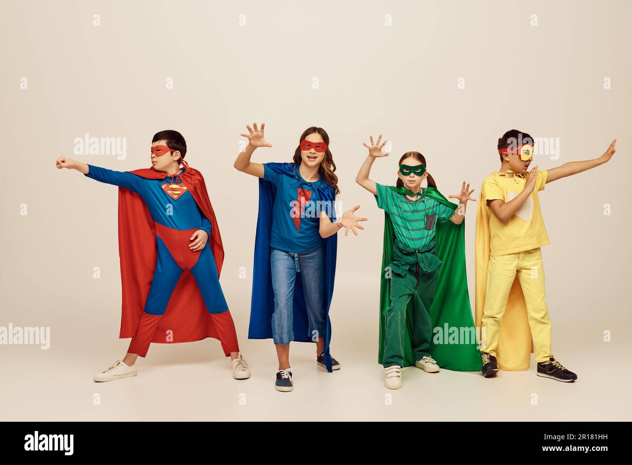 Multikulturelle Kinder in farbenfrohen Superheldenkostümen mit Umhängen und Gesten zusammen auf grauem Hintergrund im Studio, International Children's d Stockfoto