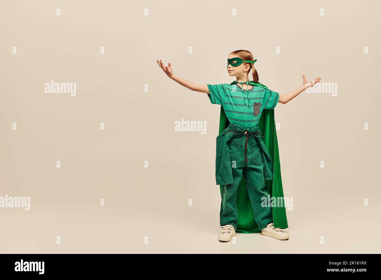 Mächtiges Mädchen in Superheldenkostüm mit grünem Umhang und Maske im Gesicht steht mit ausgestreckten Händen, zeigt Kraft und feiert Internati Stockfoto