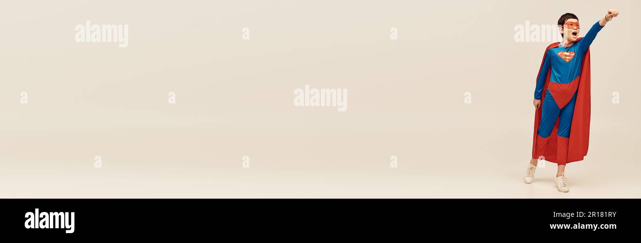 asiatischer Junge in Superheldenkostüm mit Umhang und Maske schreiend, während er Kraft zeigt, während er mit ausgestreckter Hand während des Kinderschutzes steht Stockfoto