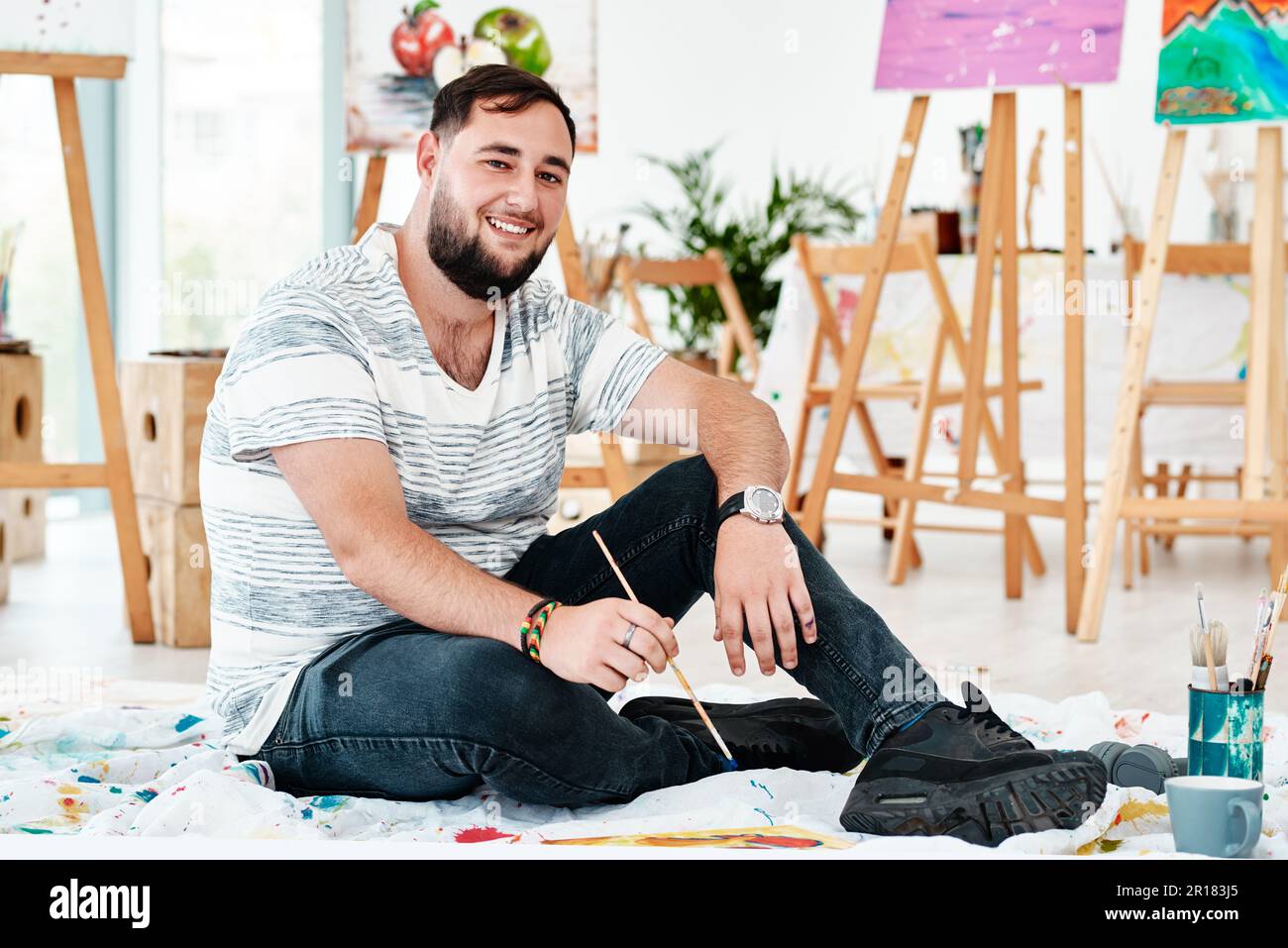 Nur ein typischer Tag in einem Kunststudio. Das ganze Porträt eines gutaussehenden jungen Künstlers, der allein sitzt und während eines Kunstkurses im Studio malt. Stockfoto